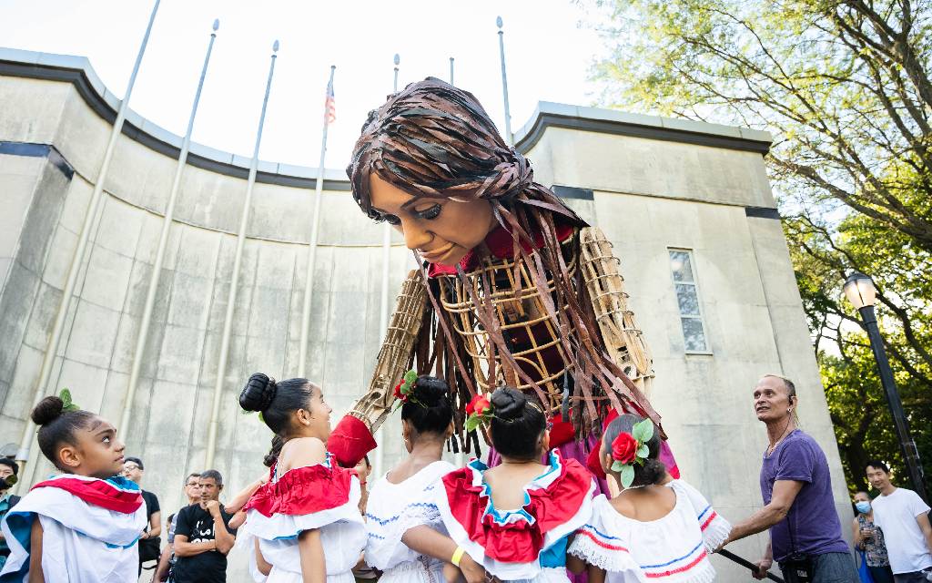 Amal la marioneta gigante en una calenda en Oaxaca, rodeada de gente, transmitiendo un mensaje de esperanza.
