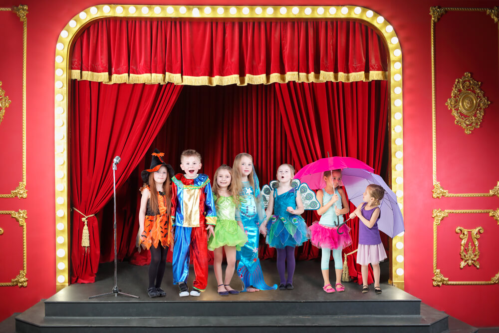 Función de teatro infantil con actores disfrazados de personajes fantásticos en escenario decorado con colores vivos frente a niños sentados en el suelo