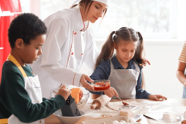 Niños con gorros de chef en taller de cocina infantil amasando pizzas sobre mesa de madera con ingredientes coloridos alrededor